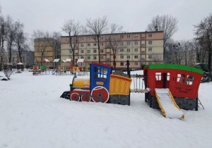 Ogród przedszkolny w zimowej szacie, nowoczesna, kolorowa lokomotywa oraz plac zabaw.
